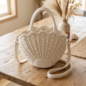 May include: A cream-colored crochet handbag shaped like a seashell, featuring a scalloped top edge and a structured base. The exterior is adorned with evenly spaced white faux pearls and includes a matching crochet handle and a detachable shoulder strap.
