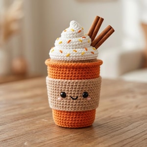 May include: A crocheted orange cup with a tan sleeve featuring a smiling face with black eyes and pink cheeks. The top has white crocheted whipped cream with orange and yellow sprinkles and two brown cinnamon sticks protruding from the top.