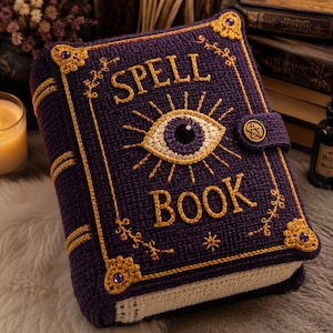 May include: A purple crocheted book cover featuring the text SPELL BOOK in yellow yarn. The cover displays a central eye motif with yellow rays, gold corner accents with purple gems, and a side strap secured by a circular gold pentagram button.