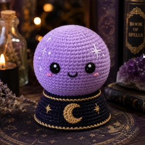May include: A round, lavender crochet plush toy shaped like a crystal ball with a smiling face, black eyes, and pink cheeks. It features white embroidered stars and sits on a dark blue base decorated with a gold crescent moon and stars.
