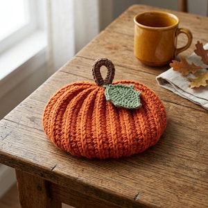 May include: A round, orange crocheted pumpkin featuring a brown crocheted stem and a single textured green crocheted leaf attached to the top. The pumpkin has a ribbed, yarn-stitched surface texture and is crafted from soft, thick orange yarn.