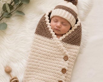 May include: A crochet baby swaddle blanket and matching beanie hat set. The swaddle is cream-colored with a brown top section and three brown wooden buttons. The beanie is cream and brown striped with a brown pom-pom on top.