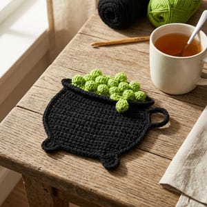 May include: A black crochet coaster shaped like a cauldron, featuring a cluster of small, bright green crocheted spheres emerging from the top. The coaster has a flat, textured surface with a small handle on the right side and three short feet.