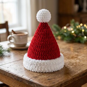Puede incluir: Un gorro de Papá Noel cónico hecho de hilo rojo con un borde grueso y difuso de color blanco y un pompón blanco a juego en la parte superior. La textura del hilo rojo es claramente visible como una serie de bucles interconectados.