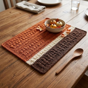 May include: A rectangular crochet placemat featuring horizontal color-blocked stripes in burnt orange, cream, and dark brown. The textured yarn pattern consists of repeating raised stitches. A wooden spoon rests on the table next to the placemat.