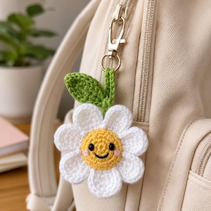 May include: A crocheted daisy keychain featuring a yellow center with a stitched black smiling face and pink cheeks, surrounded by white petals and a green leaf. The flower is attached to a silver-toned metal lobster clasp and ring.
