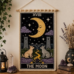 Peut inclure: Une tenture murale rectangulaire en crochet représentant la carte de tarot La Lune. Le motif présente un croissant de lune jaune avec un visage, deux tours, un chien, un loup et un chemin sinueux sur fond noir. Le texte XVIII et THE MOON est visible.