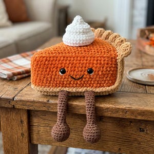 Puede incluir: Un peluche de ganchillo con forma de porción de tarta de calabaza con corteza tostada, relleno naranja y un copete de nata montada blanca. La porción tiene dos ojos negros de seguridad, una sonrisa bordada y dos piernas marrones colgantes.