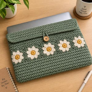 May include: A sage green crocheted laptop sleeve featuring a row of five white and yellow daisy motifs across the front flap. The sleeve is secured with a single wooden button closure and contains a silver laptop inside the protective pouch.