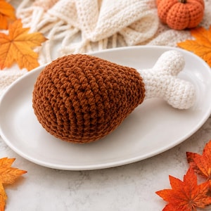 Pode incluir: Um brinquedo de coxa de frango em crochê, com um corpo de fio castanho texturizado e uma extremidade de fio branco que representa o osso. O item de pelúcia tem a forma de uma perna de ave cozinhada, com uma estrutura arredondada e firme.