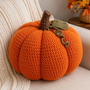 May include: A round, orange crochet pumpkin pillow featuring a textured, ribbed surface. The pumpkin is topped with a dark brown crocheted stem and a single, detailed olive green crocheted leaf with a small, curly vine attached to the top.