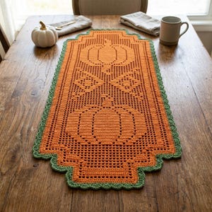 Pode incluir: Um caminho de mesa retangular de crochê laranja com um motivo de abóbora e uma borda verde festonada. O intrincado design vazado exibe duas abóboras centralizadas no tecido, criando um padrão texturizado em toda a peça.