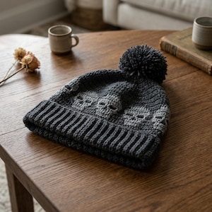 May include: A dark charcoal gray crocheted beanie featuring a repeating pattern of light gray skulls around the crown. The hat includes a ribbed brim and a large, matching dark gray pom-pom attached to the top center of the knit headwear.