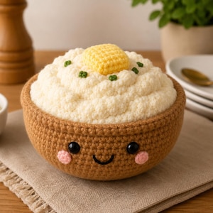 May include: A crocheted bowl of white mashed potatoes topped with a yellow pat of butter and green chives. The brown bowl features a stitched smiling face with black eyes and pink cheeks, crafted from yarn with a textured, soft appearance.