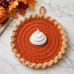 May include: A round, crocheted pot holder shaped like a pumpkin pie with a dark orange center, a light tan scalloped crust edge, and a small loop at the top for hanging. A white, spiraled, crocheted dollop of whipped cream sits in the center.
