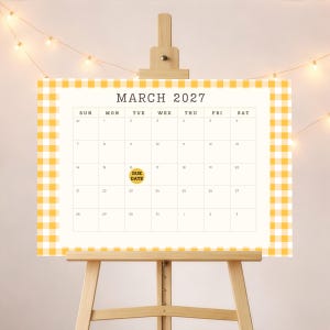 May include: A March 2027 calendar page featuring a yellow and white gingham border. The calendar grid displays days of the week and dates, with a yellow circular sticker on the 16th containing the black text DUE DATE, all displayed on a wooden easel.