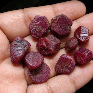 May include: A collection of ten raw, unpolished ruby gemstones with deep red coloration and irregular, multifaceted surfaces. These natural stones exhibit varying shapes and textures, showcasing the authentic, untreated appearance of the mineral.