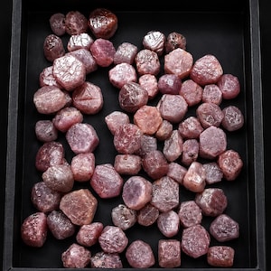 May include: A collection of numerous raw, unpolished pink and reddish-purple gemstones with irregular, multifaceted shapes. These stones are scattered across the interior of a rectangular black tray, displaying varied textures and translucent surfaces.