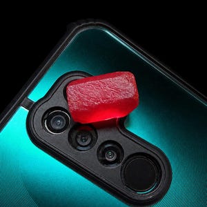 Peut inclure: Une pierre précieuse rouge translucide et rectangulaire repose sur le module photo noir d'un smartphone bleu sarcelle. Le smartphone présente une finition métallique avec un motif circulaire, tandis que le bloc photo contient trois objectifs visibles.