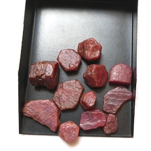 Puede incluir: Una colección de piedras preciosas de rubí en bruto de color rojo intenso con superficies irregulares y facetadas de formas variadas, que descansan dentro de una bandeja de metal negro. Las piedras muestran texturas naturales.