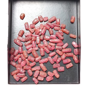 May include: A collection of small, raw, pinkish-red gemstone crystals with irregular, elongated shapes and textured surfaces. These translucent stones are scattered across a flat, rectangular black tray with raised edges.
