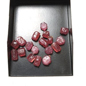 Peut inclure: Une collection de petites pierres précieuses translucides, allant du rose profond au rouge, aux formes irrégulières et facettées, dispersées sur un plateau rectangulaire en métal noir lisse. Les pierres présentent des textures variées.