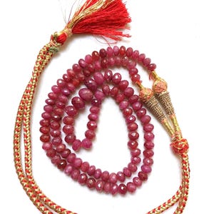 Peut inclure: Un collier composé de perles de rubis rose foncé à facettes disposées en cercle, reliées à un cordon tressé rouge et doré. Le cordon présente des embouts métalliques dorés et un pompon rouge avec des fils dorés sur le dessus.