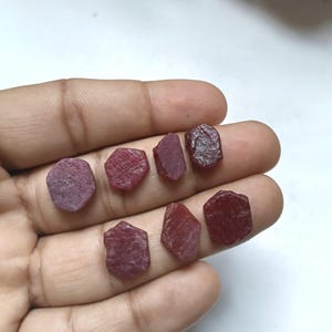 Peut inclure: Sept petites pierres précieuses rubis brutes et non polies, aux formes irrégulières rappelant l'hexagone, présentant des nuances variées de rouge profond et cramoisi. Les pierres ont une surface mate et texturée avec des inclusions naturelles.