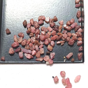 May include: A collection of small, raw, reddish-pink gemstone fragments scattered across a black rectangular tray, with several additional loose pieces resting on the white surface below the tray, showing varied shapes and translucent textures.