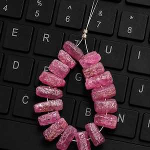Può includere: Una fila di perline di rubino rosa grezzo, di forma cilindrica, disposte a goccia. Ogni perla presenta una superficie ruvida e testurizzata con inclusioni naturali e una vivace tonalità magenta, infilate su un sottile filo metallico argentato.