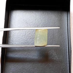 May include: A small, rectangular, translucent pale green gemstone held by a pair of silver-toned metal tweezers. The stone has a smooth, polished surface with subtle internal variations and a soft, muted colour profile, captured in a clear, focused view.