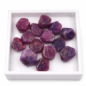 Peut inclure: Une collection de rubis bruts et non polis dans des tons variés de magenta profond, de violet et de rouge foncé. Ces pierres naturelles présentent des surfaces irrégulières et facettées, présentées dans un plateau carré blanc.