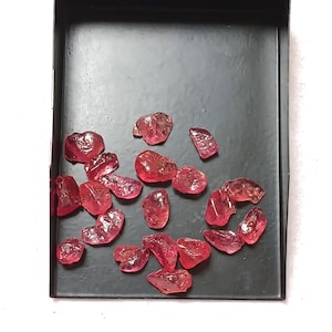 Peut inclure: Une collection de petites pierres précieuses brutes, translucides et rose-rouge, dispersées sur un plateau rectangulaire en métal noir. Les pierres présentent des formes irrégulières et dentelées avec des surfaces facettées.