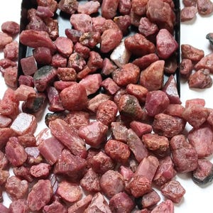 Könnte beinhalten: Eine Sammlung roher, unpolierter Rubinedelsteine in verschiedenen Rosa- und Dunkelrottönen. Die Steine haben unregelmäßige Formen und Texturen, wobei einige durchscheinend und andere undurchsichtig wirken, angehäuft in einer dunklen Schale.