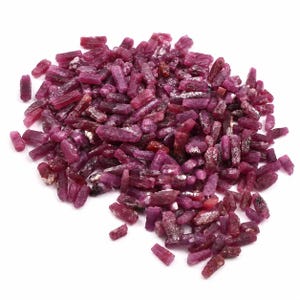 Può includere: Una collezione di piccoli cristalli di rubino grezzi e non lucidati in varie tonalità di magenta intenso e rosso scuro. Questi frammenti di pietre preziose naturali presentano forme prismatiche allungate con superfici ruvide e strutturate.
