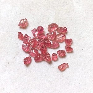Op de afbeelding: Een verzameling kleine, ruwe, ongepolijste roze-rode edelsteenfragmenten met onregelmatige, hoekige vormen en doorschijnende oppervlakken. Deze losse, natuurlijke mineraalstukken vertonen variërende kleurdieptes en kristallijne texturen.