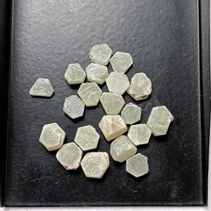May include: A collection of pale green, translucent, hexagonal-shaped mineral stones with visible internal crystalline structures and varying surface textures, scattered across a flat, solid black surface.