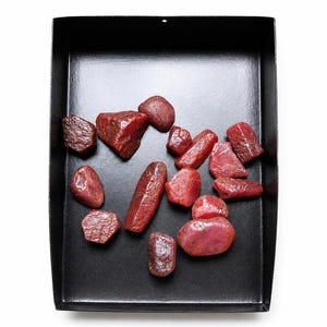 Puede incluir: Una colección de piedras preciosas de rubí en bruto y sin pulir esparcidas por el interior de una bandeja rectangular de metal negro. Las piedras varían en tamaño y forma, mostrando texturas naturales, tonos carmesí y superficies opacas.