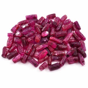 May include: A collection of numerous small, elongated, cylindrical ruby gemstones. Each piece displays a vibrant, deep magenta-red hue with natural surface textures, internal inclusions, and varying degrees of translucency throughout the pile.