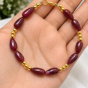 May include: A bracelet featuring alternating dark red, teardrop-shaped gemstone beads and small, round, polished gold-tone metal beads. The piece is secured with a gold-tone metal lobster claw clasp, creating a continuous circular design.
