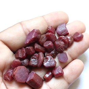 Puede incluir: Una colección de pequeñas piedras preciosas de rubí en bruto, sin cortar, con un color rojo intenso y formas irregulares y facetadas. Estas piedras naturales presentan texturas variadas, desde superficies lisas hasta bordes rugosos y mates.