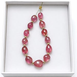 Puede incluir: Un collar de cuentas de turmalina rosa pulidas y translúcidas, intercaladas con pequeños espaciadores metálicos de tono dorado. Las cuentas tienen formas irregulares y tamaños variados, dispuestas en un bucle y aseguradas con un hilo dorado.