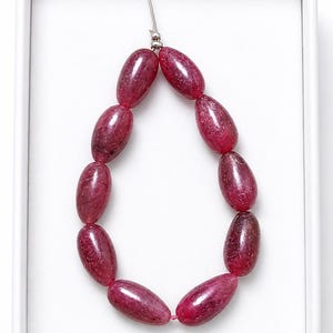 May include: A strand of ten polished, teardrop-shaped beads in a deep magenta-red hue with natural internal inclusions. The beads are strung on a thin cord and arranged in an oval loop, secured at the top with a small silver-toned metal bead.