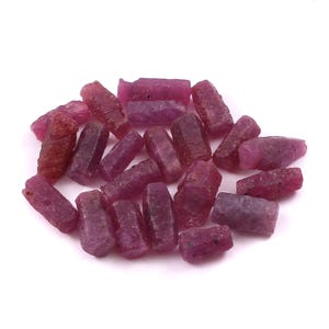 Può includere: Una collezione di piccoli cristalli di rubino grezzi e non lucidati con una tonalità magenta profonda e rosso rosato. Questi pezzi di pietre preziose naturali presentano forme esagonali allungate e irregolari con superfici opache.