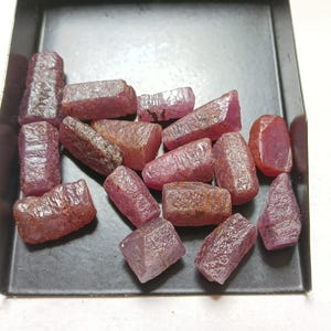 Peut inclure: Une collection de rubis bruts et non polis dans des nuances variées de rose profond et de rouge. Ces pierres présentent des formes naturelles irrégulières avec des surfaces cristallines texturées, disposées dans un plateau en métal noir.