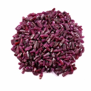 Op de afbeelding: Een dichte hoop kleine, ruwe robijn-edelsteenfragmenten. De stukjes zijn onregelmatig van vorm en grootte en hebben een diepe, verzadigde magenta-rode kleur met een natuurlijke, ongepolijste textuur en variërende gradaties van doorschijnendheid.