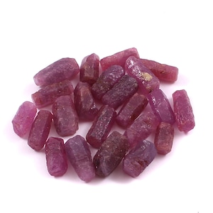Könnte beinhalten: Eine Ansammlung kleiner, roher, unpolierter Rubinkristalle in verschiedenen Schattierungen von tiefem Pink und Rot. Jeder Stein weist eine natürliche, längliche hexagonale Form mit rauer, texturierter Oberfläche und halbtransparenter Beschaffenheit auf.