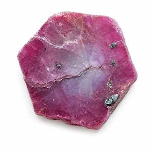 Puede incluir: Una rodaja de rubí de forma hexagonal plana con un color magenta vibrante y rosa intenso. La superficie muestra texturas cristalinas naturales, fracturas internas y pequeñas inclusiones minerales metálicas oscuras en la piedra.