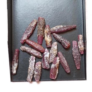 May include: A collection of elongated, raw ruby crystals scattered on a black tray. Each crystal features a deep magenta to reddish-purple hue with rough, textured surfaces and visible mineral inclusions, displaying natural, unpolished formations.