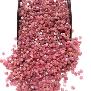 Op de afbeelding: Een grote verzameling kleine, ruwe, ongepolijste roze-rode edelsteenfragmenten die uit een zwarte metalen schep stromen. De stenen hebben onregelmatige, gefacetteerde vormen met variërende tinten van diep magenta tot lichtroze kleuren.